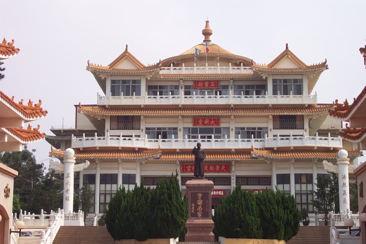 天極行宮正面全景。建築為三層閣樓式，金色琉璃瓦屋頂，各層正面懸掛紅底金字牌匾，由下至上分別書「天帝教天極行宮」、「大同堂」、「玉靈殿」。廣場中央立有涵靜老人銅像，兩側石柱分書「正大光明」、「浩然正氣」。整體建築莊嚴宏偉，融合傳統廟宇與樓閣風格。