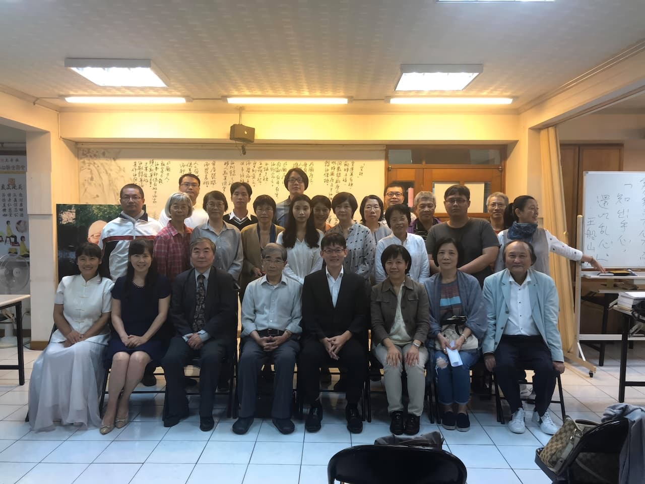 約二十五名學者於學術文化活動會後合影；室內擺有折疊椅，後方牆面掛有行書長卷，右側立有白板