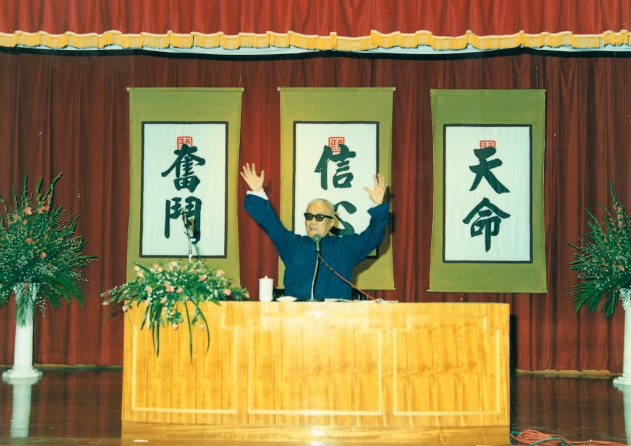 涵靜老人（李玉階先生）在演講台上慷慨演說，雙手高舉過頭，神情激昂。講台後方垂掛三幅綠底書法橫幅，由右至左書有「天命」、「信心」、「奮鬥」，配以紅色舞台布幕。台上備有麥克風與鮮花，為宗教哲學研究社公開演講系列現場。
