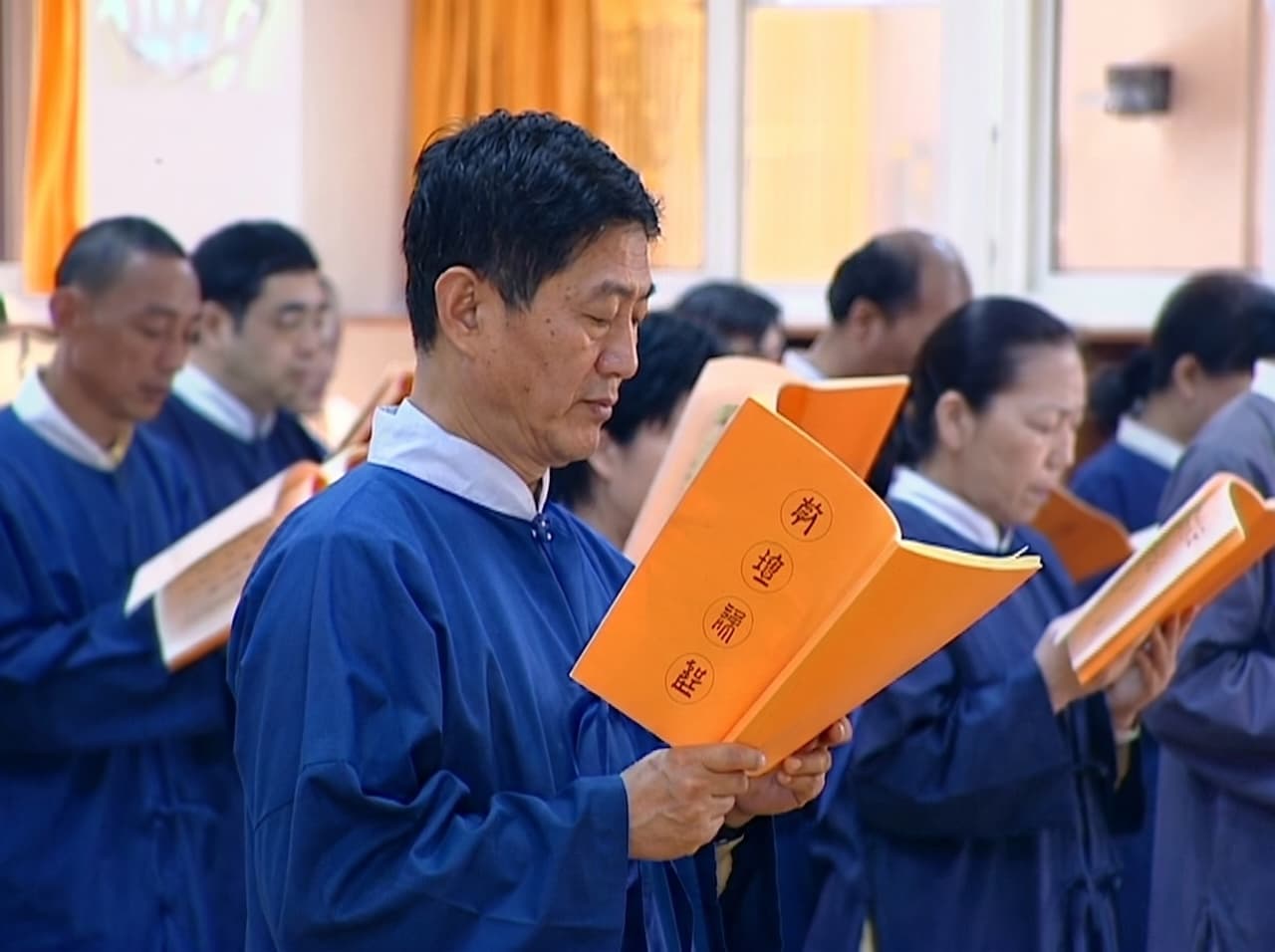 天帝教同奮集體持誦皇誥場景。前景一名男性同奮身著深藍色道袍，手持橙色封面皇誥冊，雙目低垂，神情專注虔誠；身後多排同奮同樣持冊低誦，隊列整齊，氛圍莊嚴肅穆。