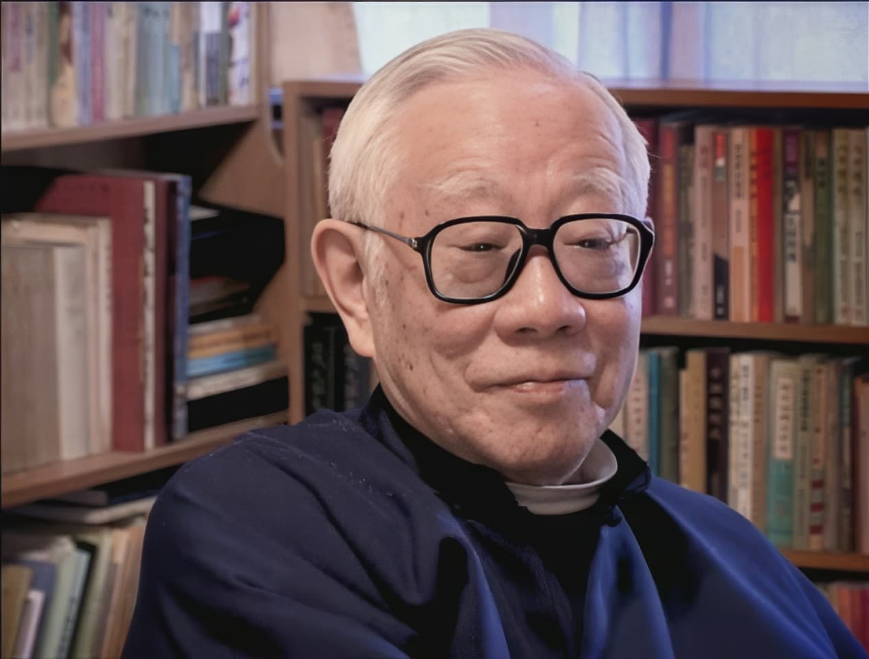 李維生首席使者半身近景肖像。老人銀白短髮，配戴深色方框眼鏡，身著深藍色中式道袍，神情慈藹沉穩，嘴角帶有微笑。背景為滿架典籍的書架，書香氣息濃厚，呈現飽學修道者的書齋風采。