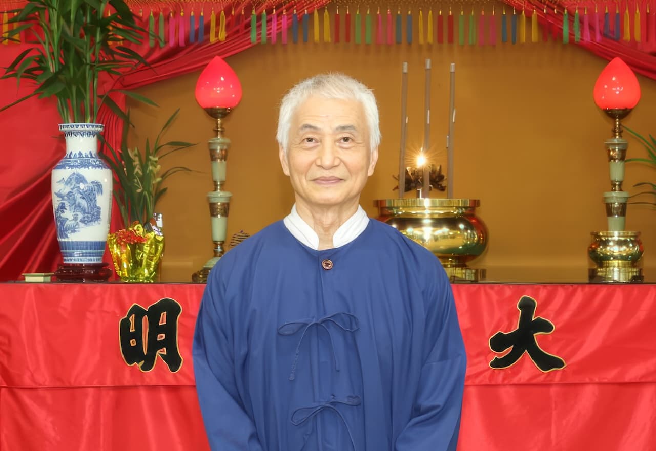 陳光理首席使者正面肖像。老人白髮，面容祥和略帶微笑，身著深藍色道袍，站立於神壇前。壇上陳列香爐、燭台、青花瓷瓶等禮器，前方紅色桌裙書有「正大光明」，因取景角度僅見「大」、「明」二字，背景掛有彩色流蘇裝飾。