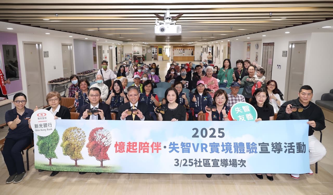 約四十名與會者於社區活動中心大廳合影，手持 VR 眼鏡；前方橫幅書「2025 憶起陪伴・失智 VR 實境體驗宣導活動 3/25 社區宣導場次」，由紅心字會與新光銀行共同主辦