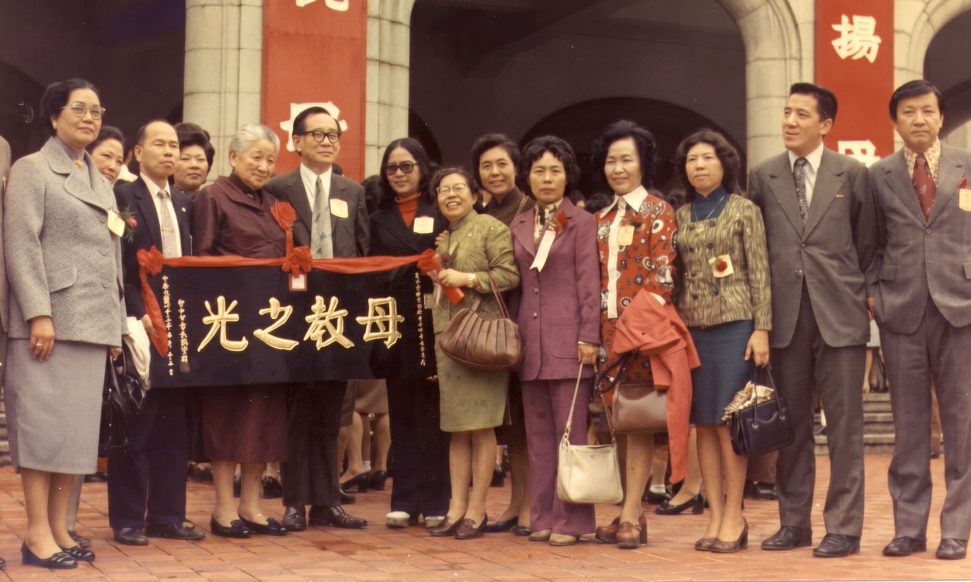 彩色照片。約十五人在一棟白色拱廊建築前排成一列，後方懸掛紅色橫幅。過純華女士著深色酒紅上衣，站於左側靠近匾額處；數人共同托舉一面深色大型匾額，匾上以金字從右至左書「母教之光」，匾額上繫紅色蝴蝶結。眾人面帶笑容。