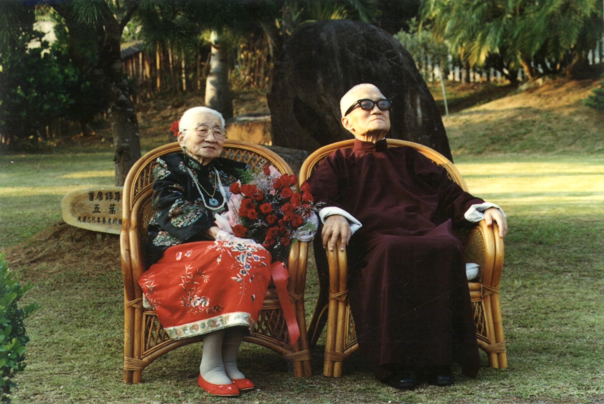 彩色照片。李玉階先生與過純華女士並肩坐於戶外藤椅上，背後有大石與棕櫚樹。過純華女士著黑色花紋上衣搭配紅色長裙，手持一束紅花，面帶笑容；李玉階先生著深酒紅色長袍，戴黑框大鏡片深色眼鏡，神情安詳。